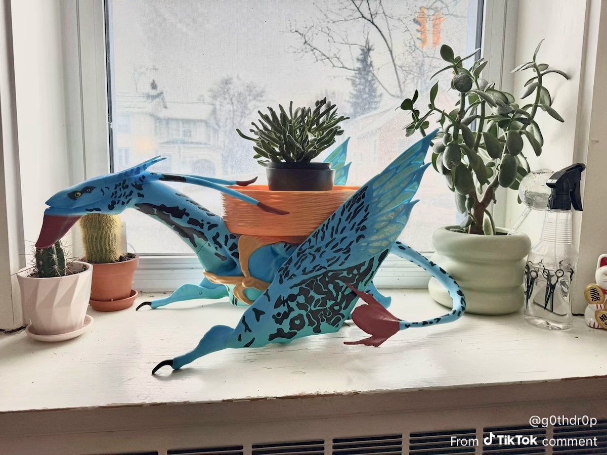 A collectible Avatar popcorn bucket, shaped like a giant blue dragon, sitting on a windowsill. It has been repurposed as a planter and has a small succulent nested in its back.