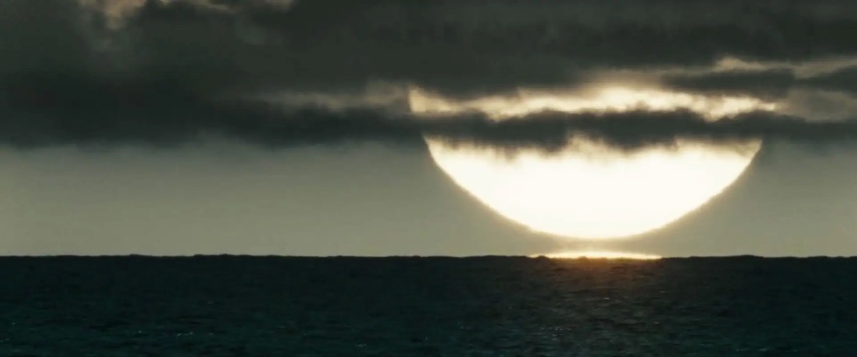 A pale sun hovers near the horizon, thin clouds across it. The water at the horizon is dark.