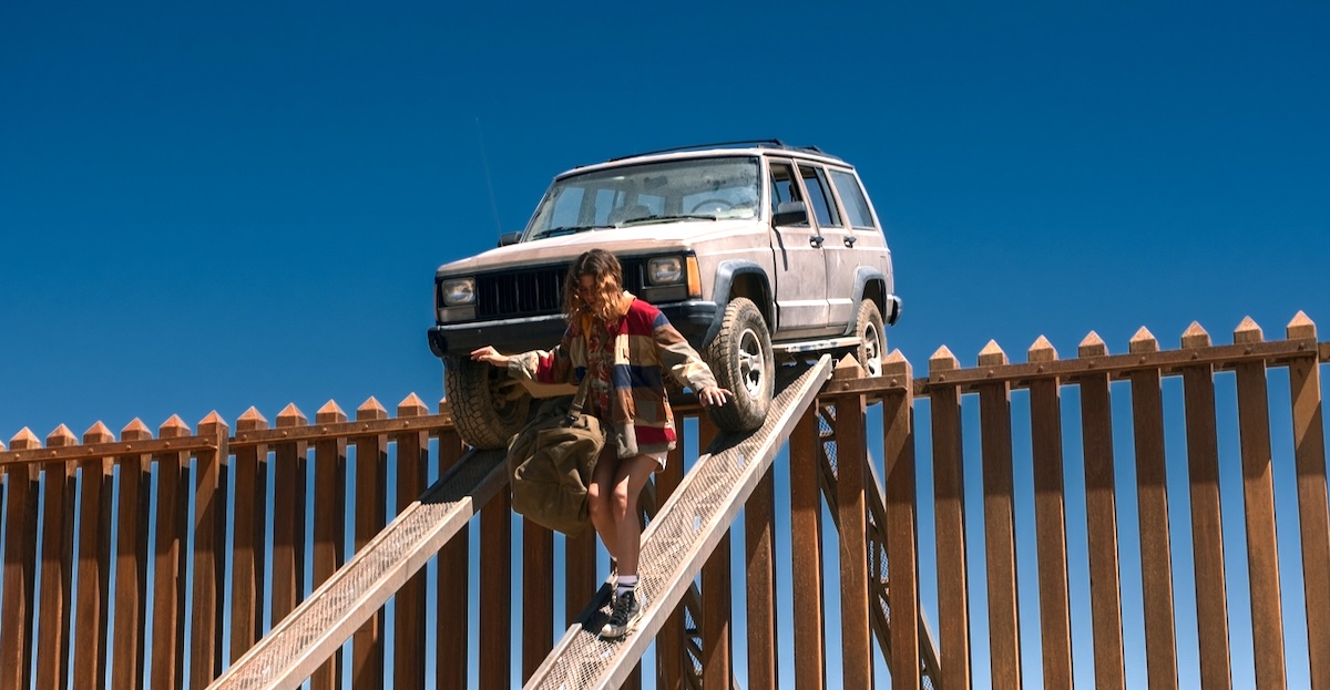 A young woman descends a wooden plank from a high metal fence, an SUV balanced precariously on the top of the fence behind her.