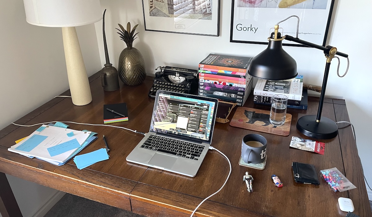 A wooden desk with a laptop on it, alongside other claptrap and paraphernalia.